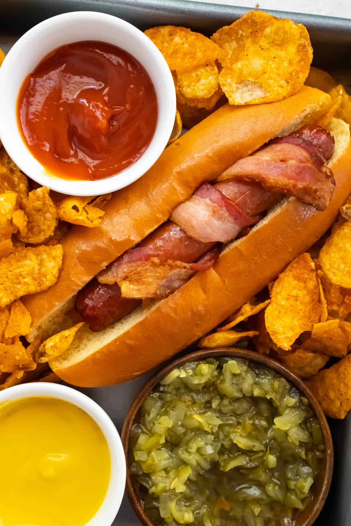 A bacon wrapped hot dog in a bun surrounded by barbecue potato chips, and small bowls of ketchup, yellow mustard, and relish.