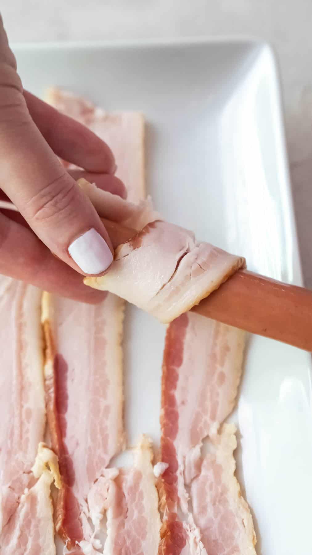 A hand wrapping a piece of bacon around a hot dog.