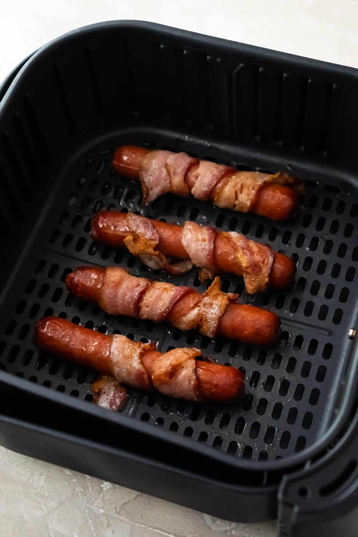 Four bacon wrapped hot dogs in an air fryer basket.
