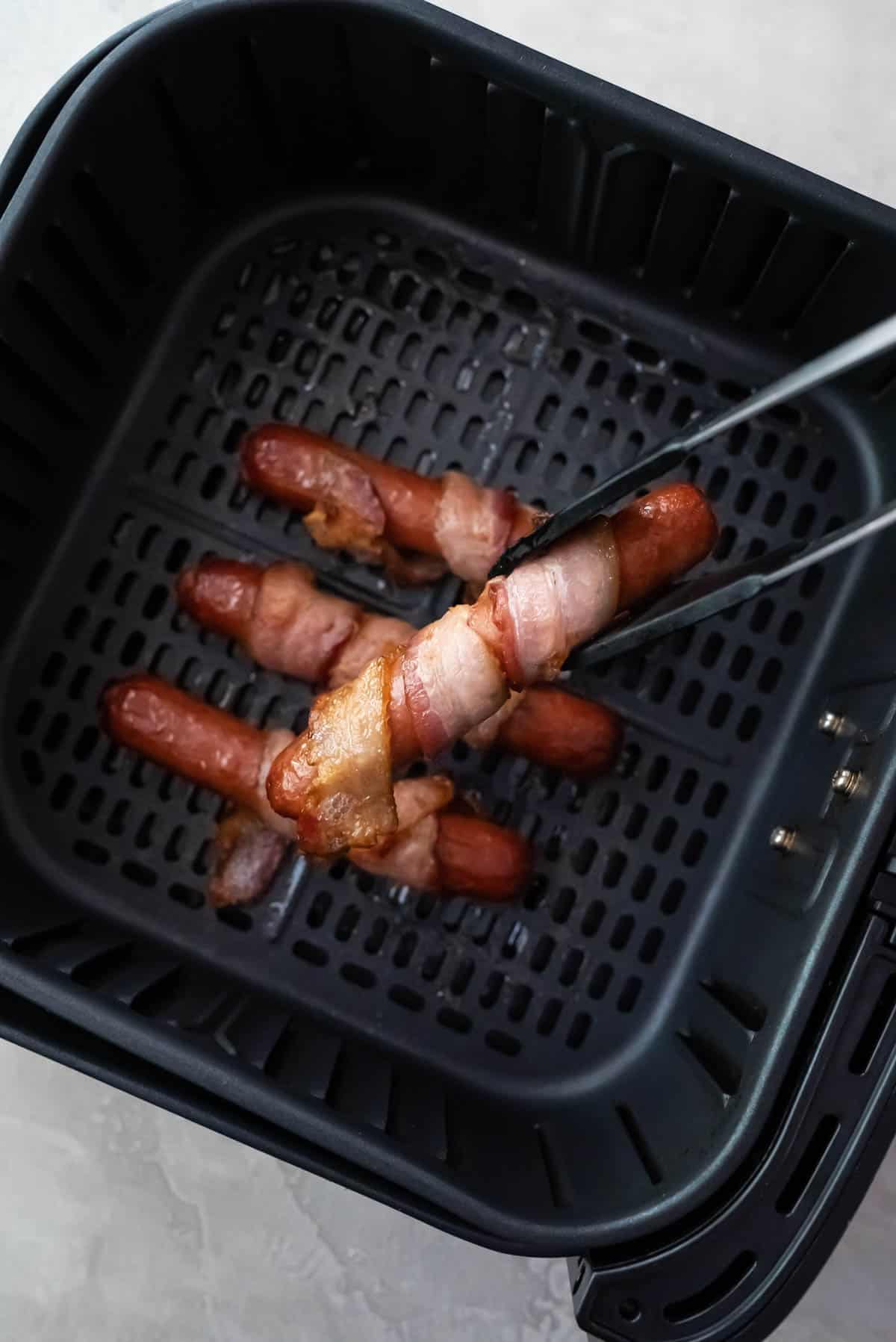 Tongs lifting a bacon wrapped hot dog out of an air fryer basket.