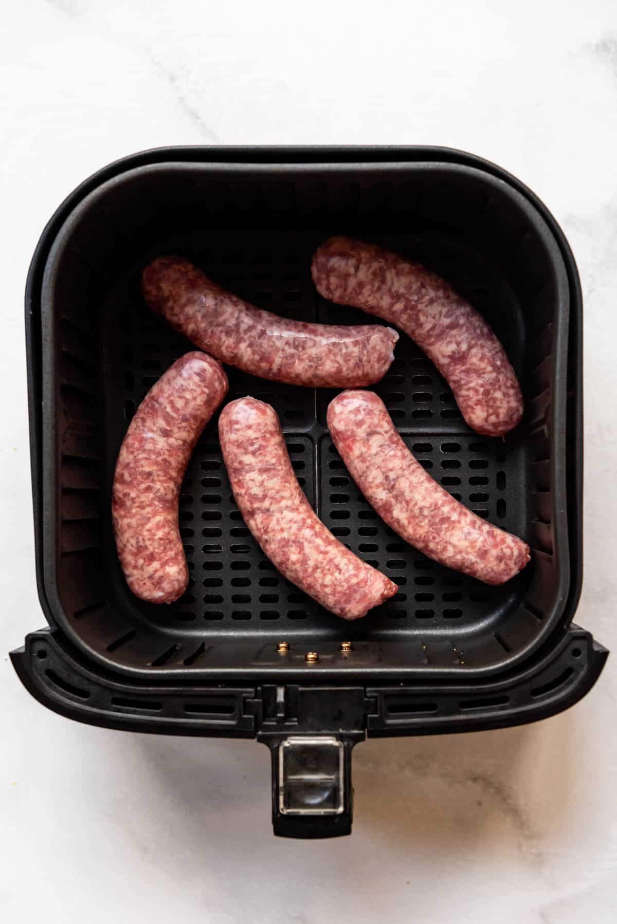 Top view of a square air fryer basket with uncooked brats sausages in it.