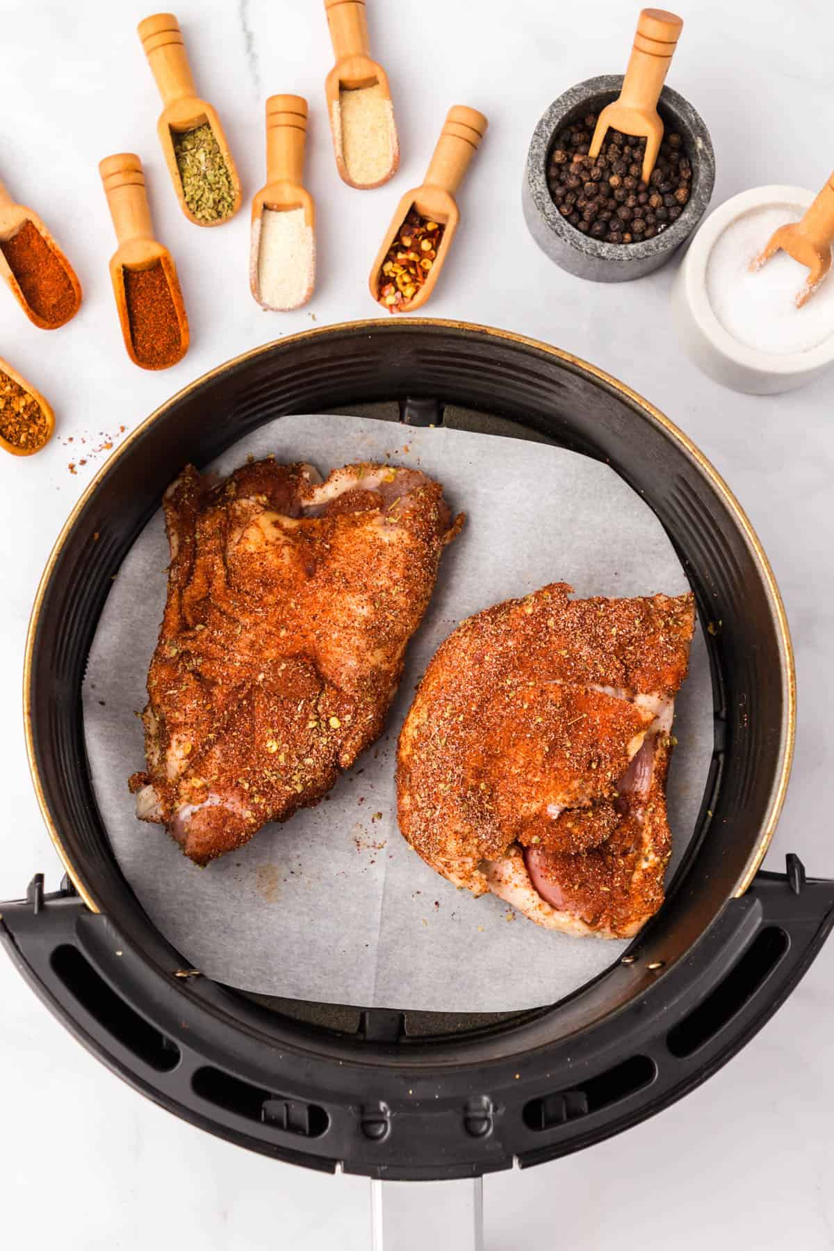 Top view of a basket of an air fryer with coated chicken thighs in it on top of parchment paper, with small spice scoops with spices in them sitting in a row above the bowl. 