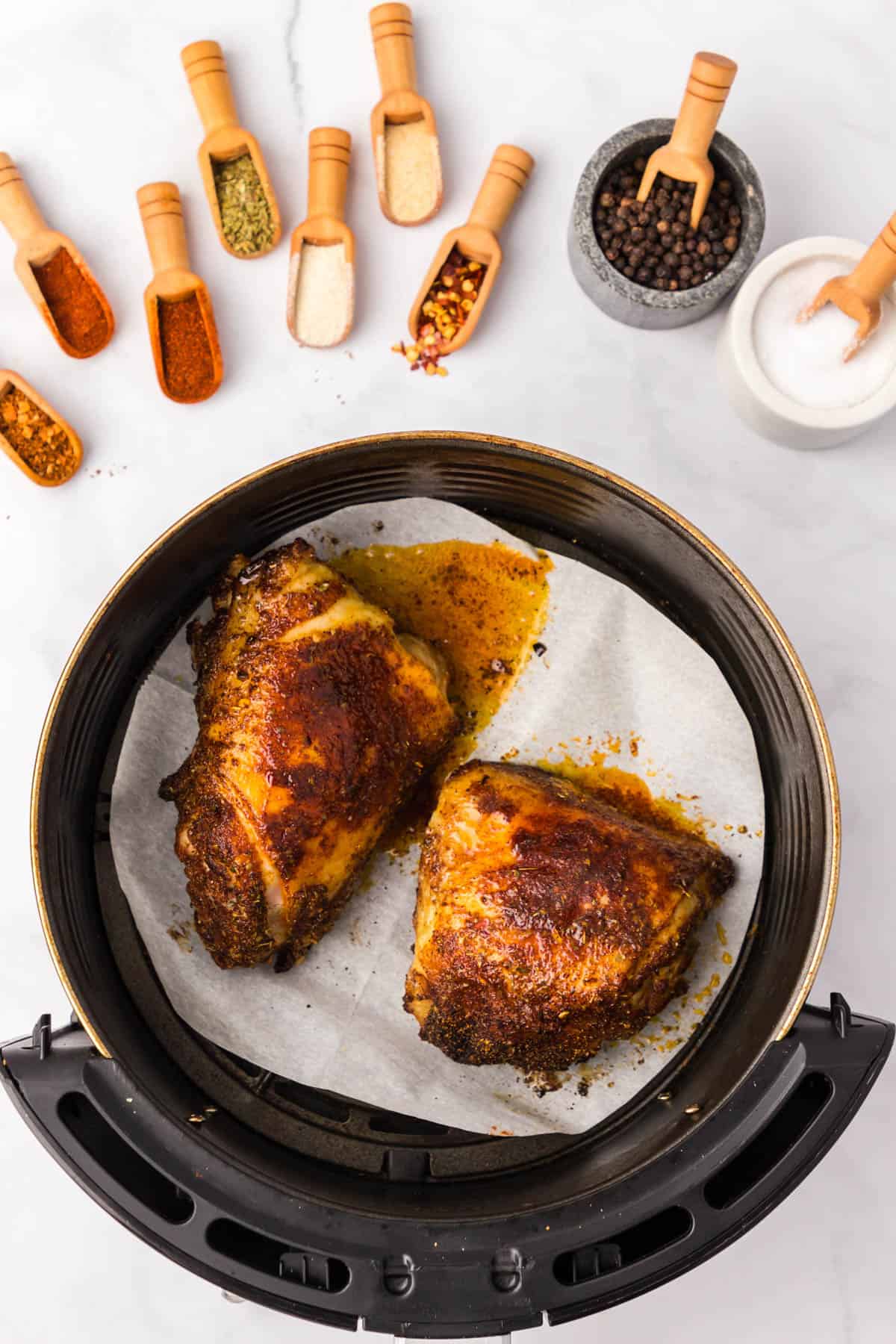 Top view of a basket of an air fryer with half cooked, coated chicken thighs in it, with small spice scoops with spices in them sitting in a row above the bowl.