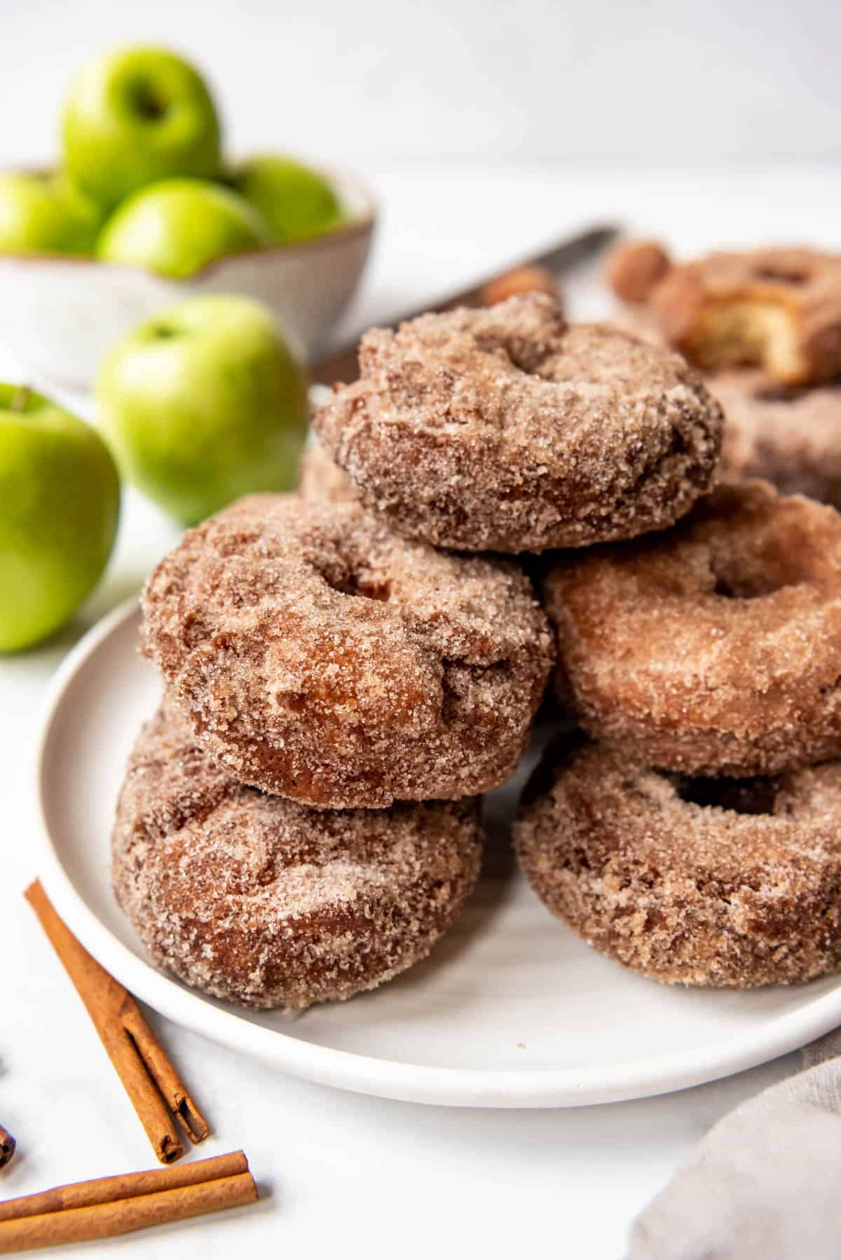 Homemade apple cider donuts stacked on a plate in front of green granny smith apples.