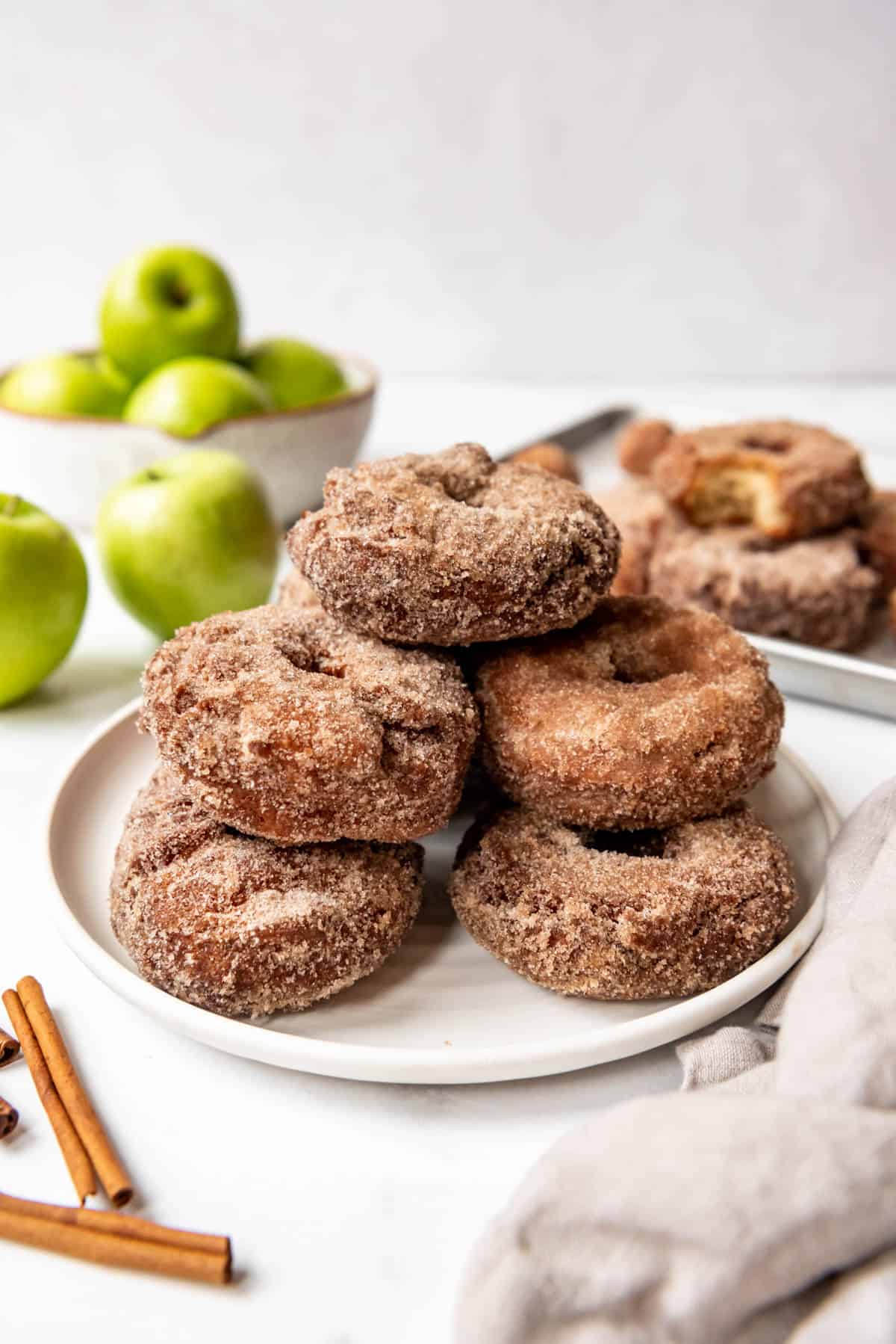 New England apple cider donuts piled on a plate with green apples behind them.