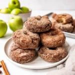 New England apple cider donuts piled on a plate with green apples behind them.