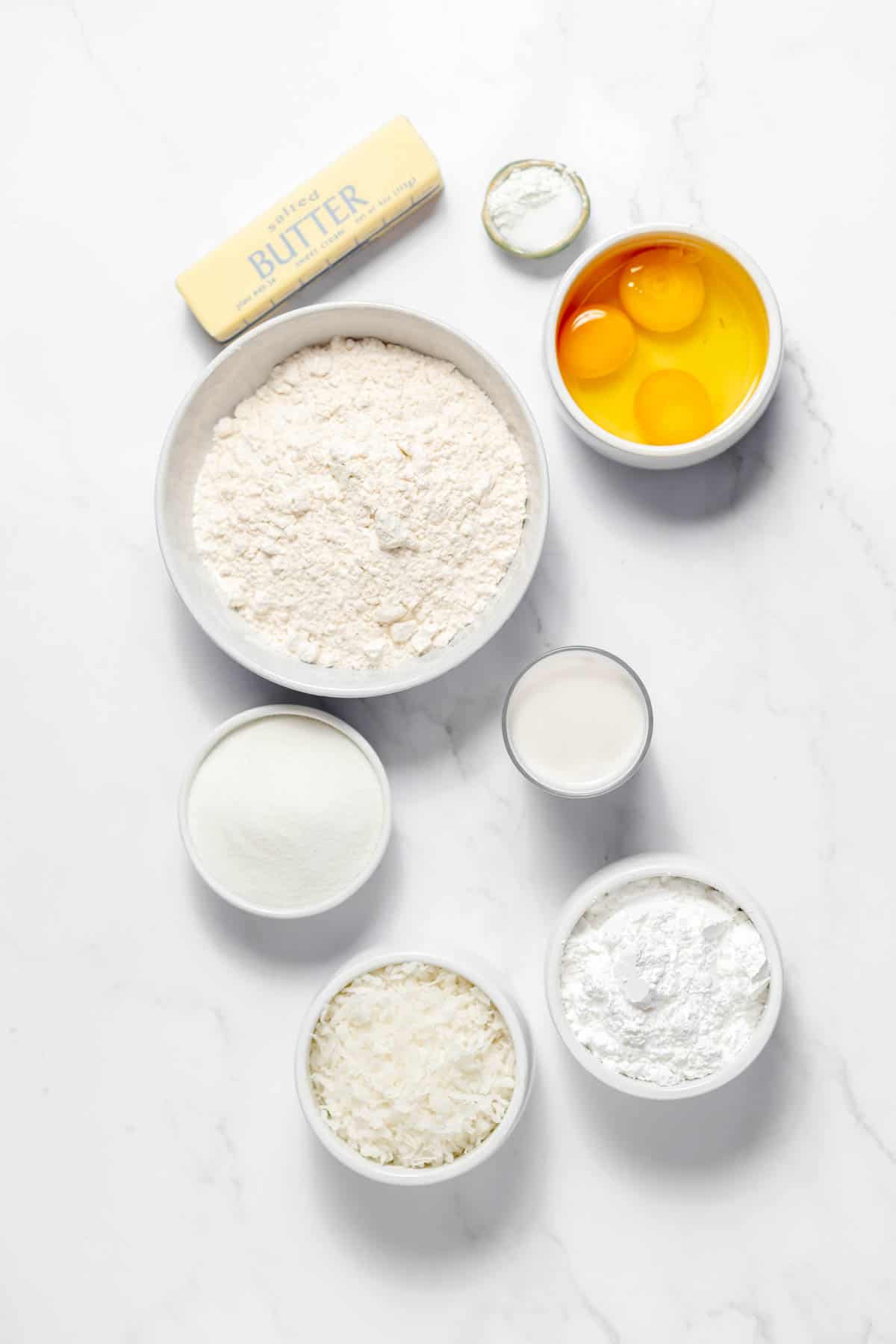 Top view of ingredients needed to make Coconut Bread in small bowls on a white surface. 