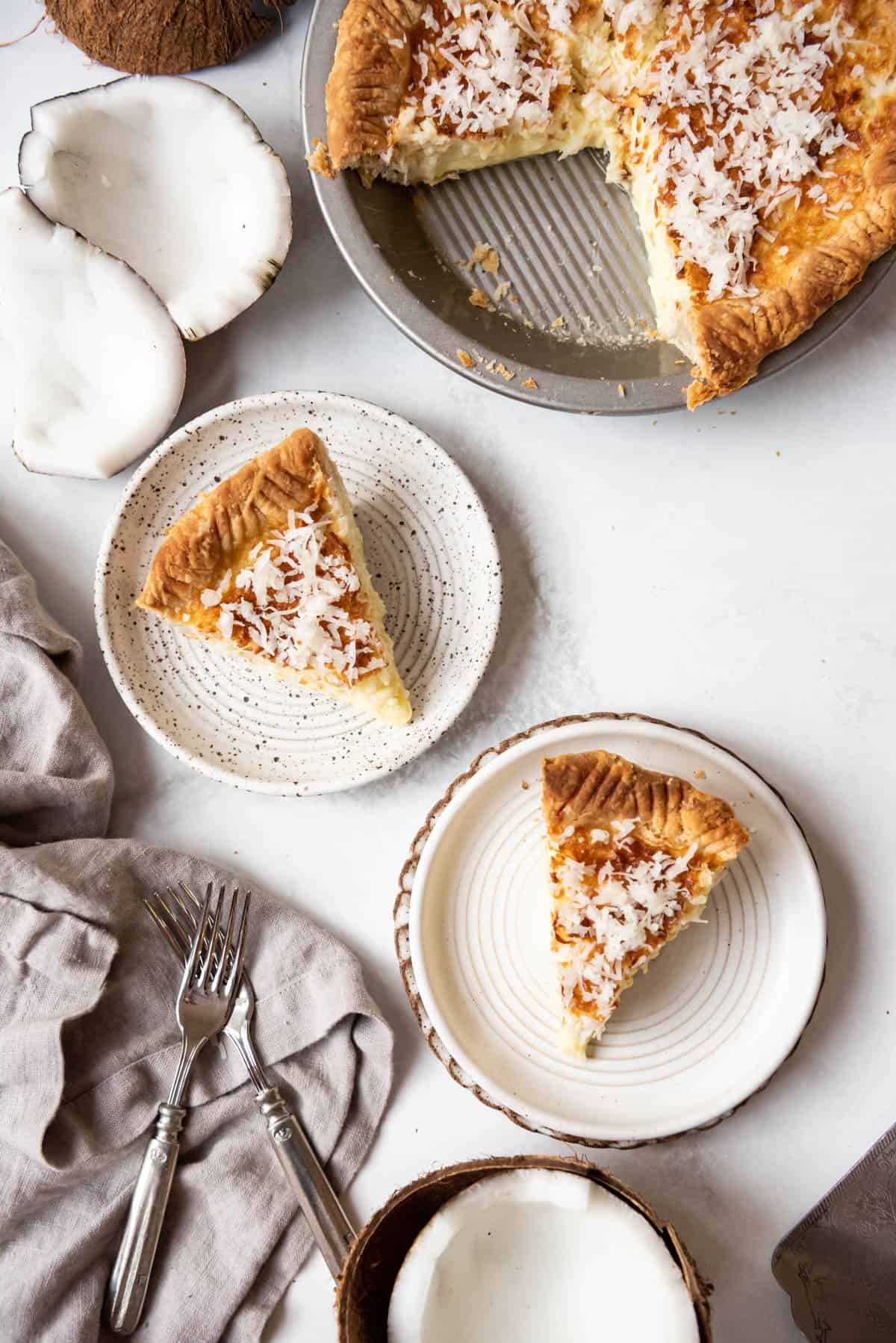 An overhead image of two slices of coconut custard pie on plates next to the rest of the pie, fresh coconut, and a cloth napkin.