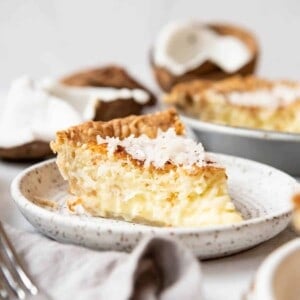 A slice of coconut custard pie on a plate with a fresh coconut behind it.