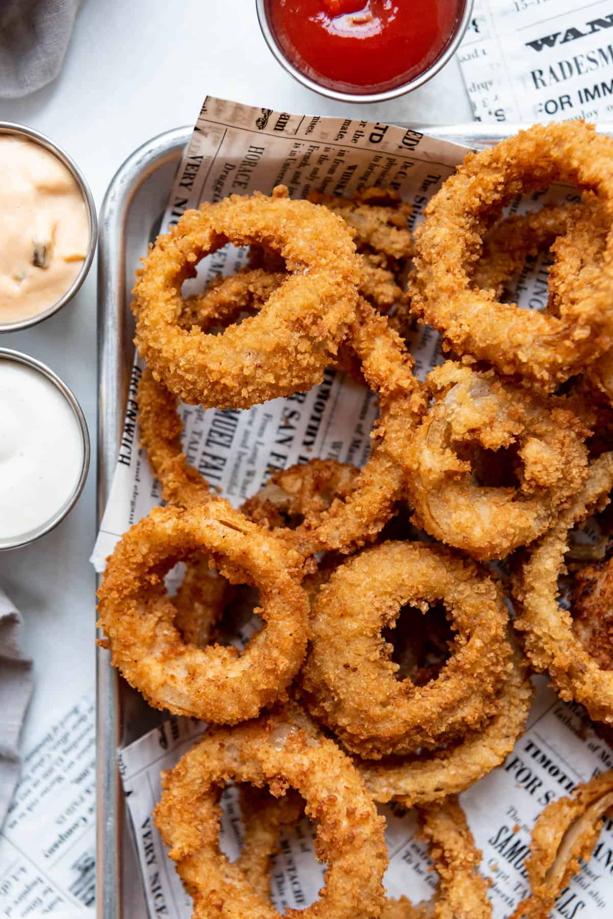 Crispy fried onion rings on newsprint next to dipping sauces.