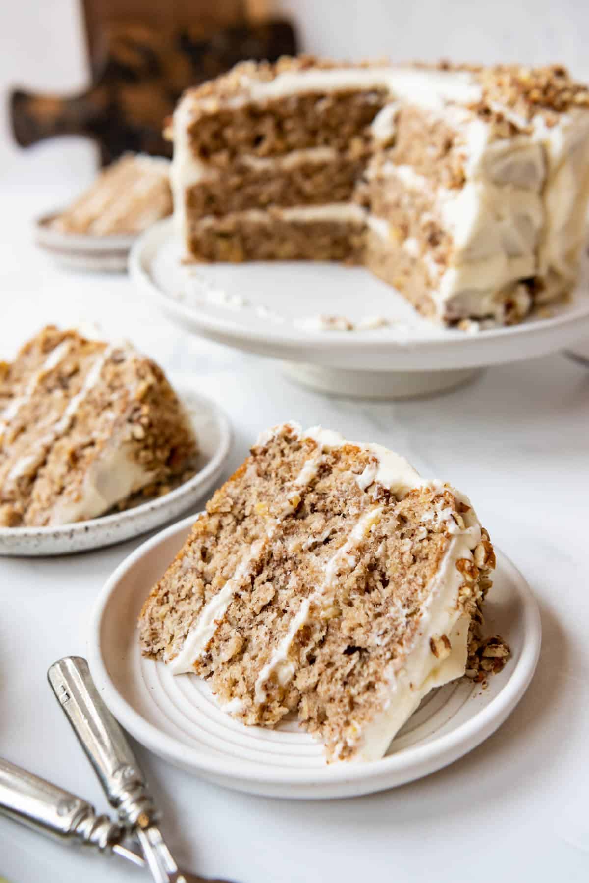 Slices of hummingbird cake on plates in front of the rest of the cake on a cake stand.