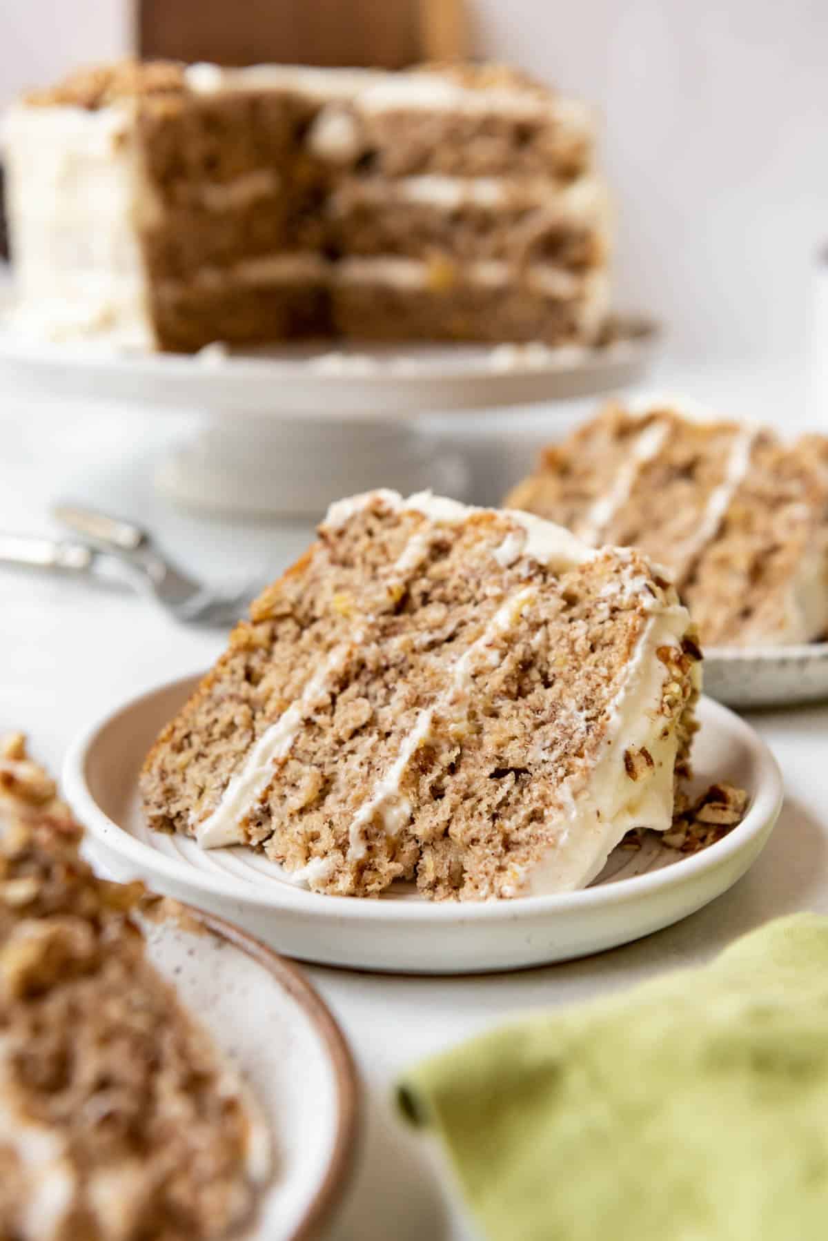 A slice of hummingbird cake on a white plate with the rest of the cake in the background and more plates of cake around it.