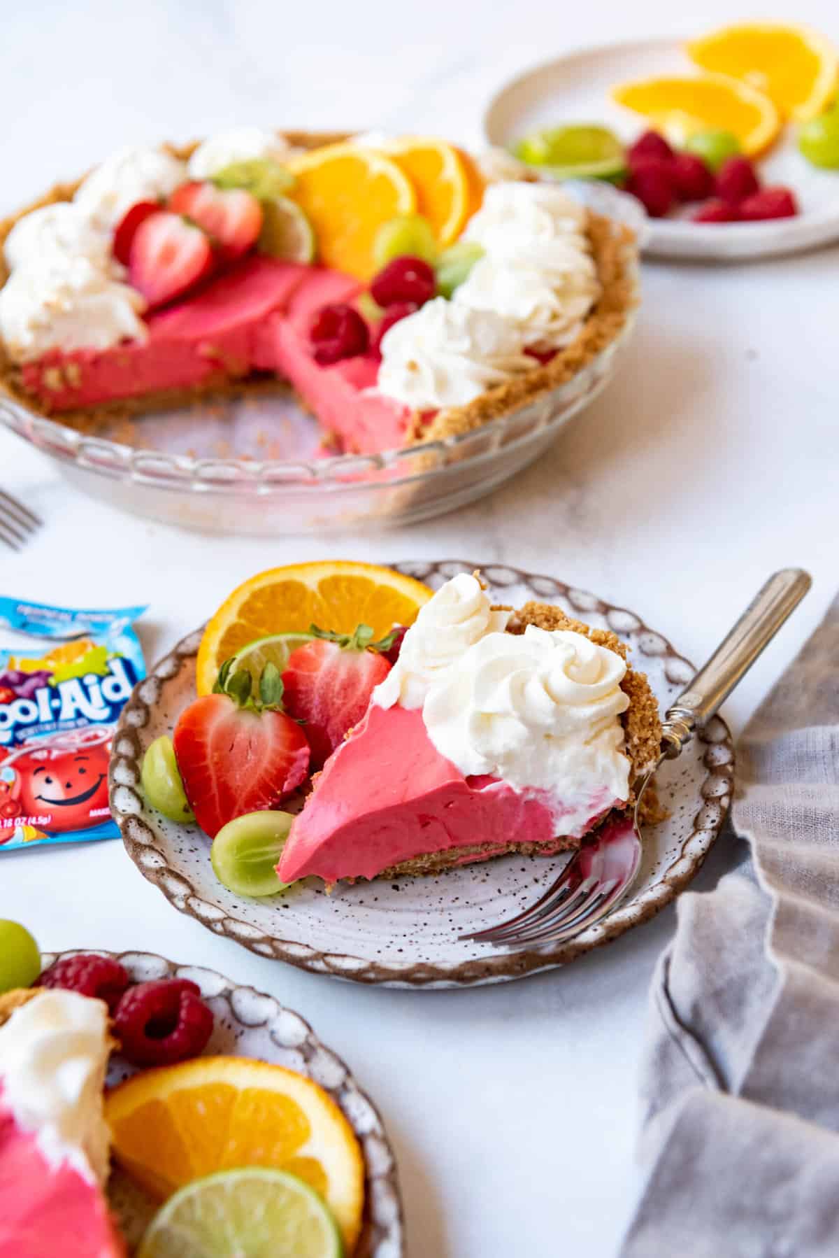 A slice of Kool-Aid pie on a plate with fresh fruit in front of the rest of the pie in a glass pie dish.