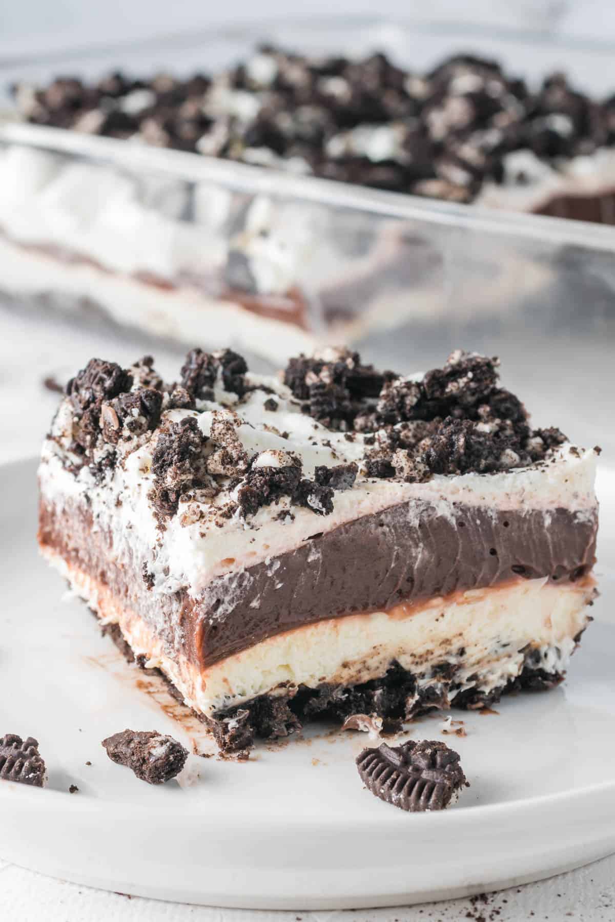 A slice of Oreo dessert on a plate in front of the rest of the dessert in a glass baking dish.