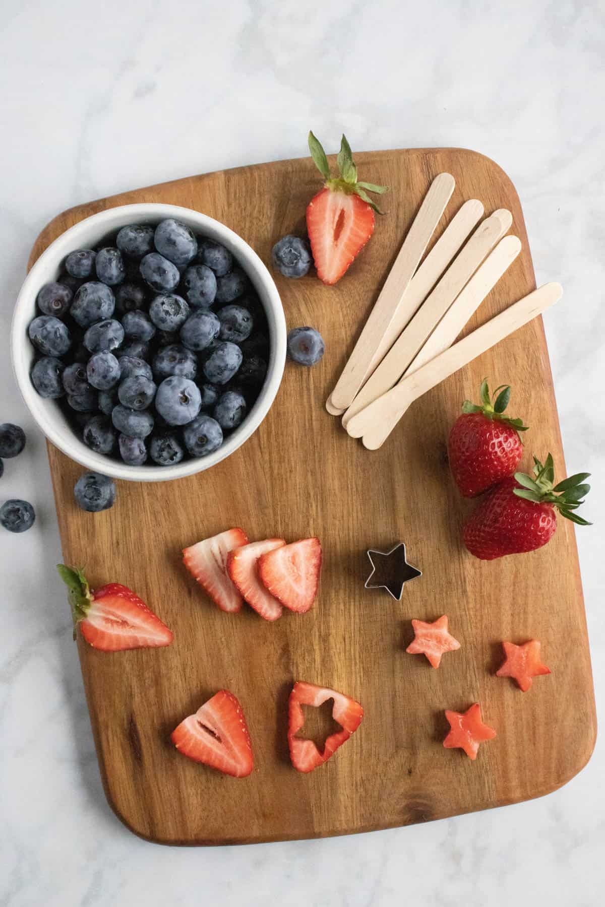 Cutting out stars from strawberry slices with a small star cookie cutter on a cutting board with popsicle sticks and blueberries in a bowl.