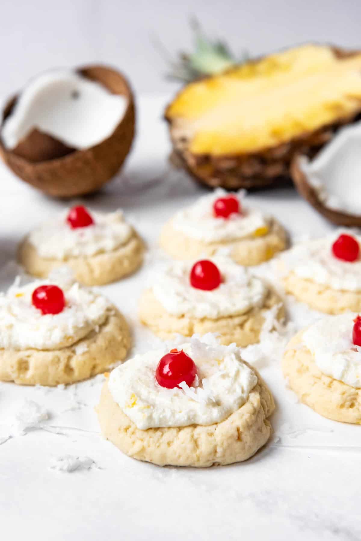 Soft pina colada cookies with frosting and maraschino cherries on top in front of a pineapple and coconut.