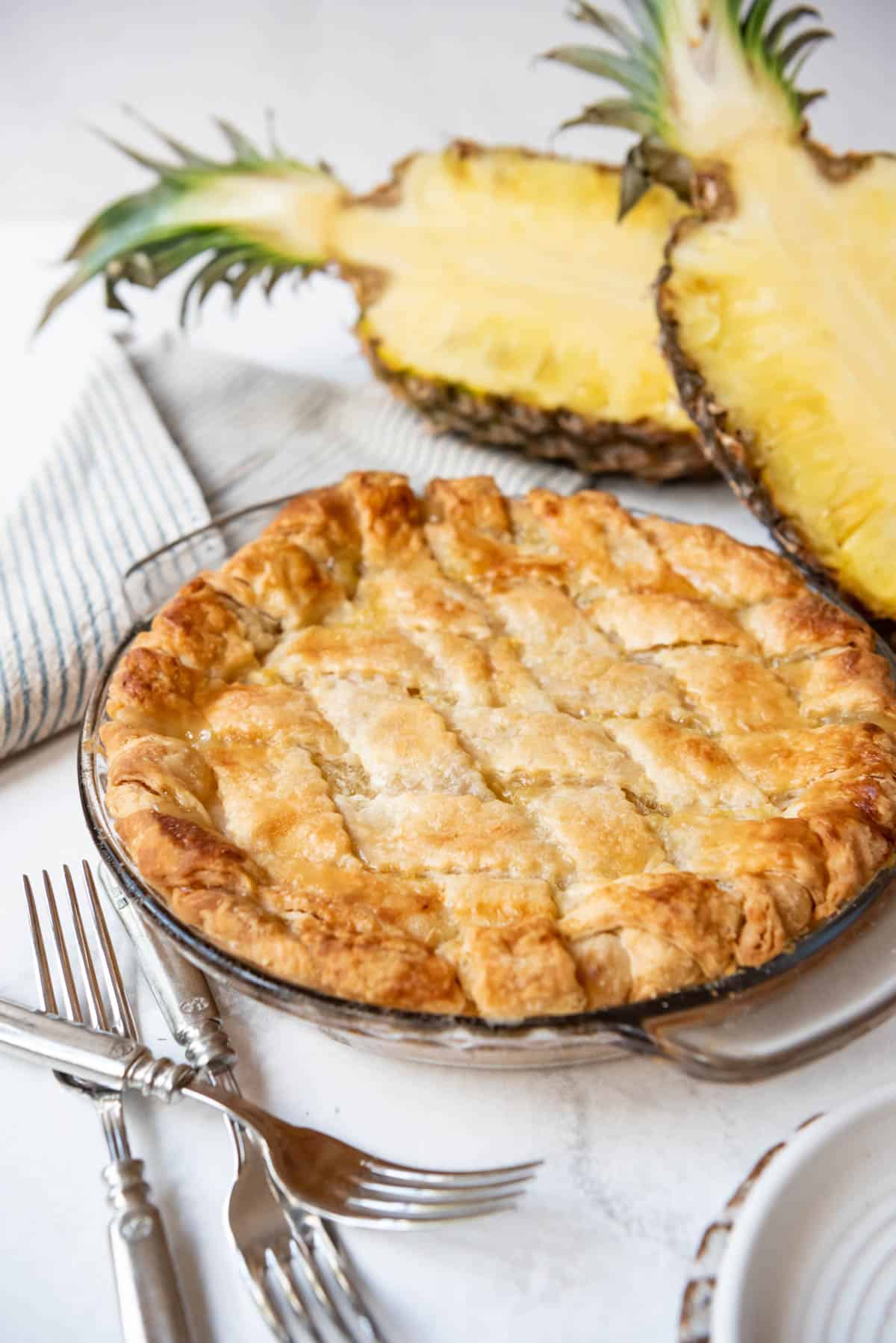 A double crust pineapple pie in a glass pie dish in front of two halves of a fresh pineapple.