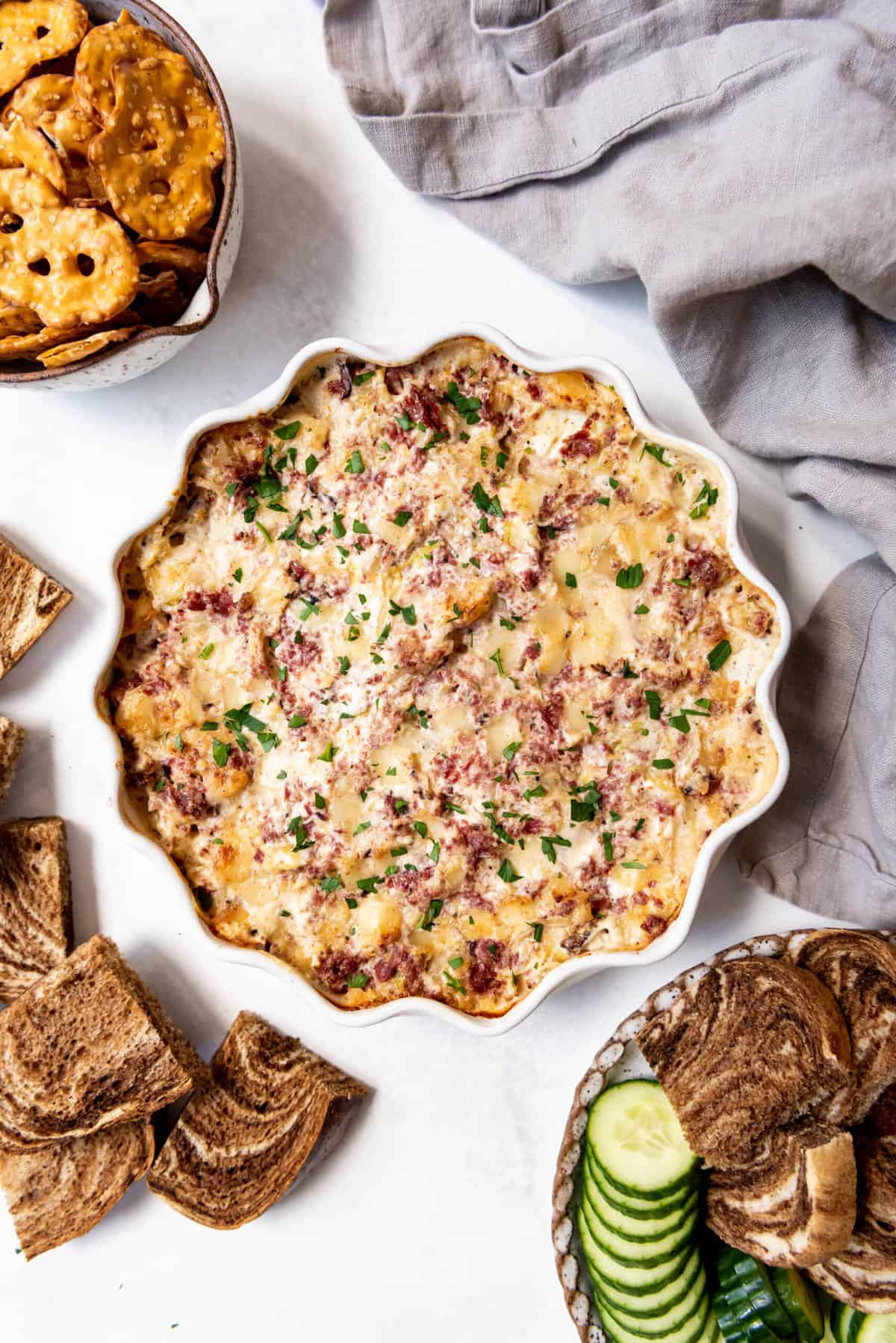 An overhead image of baked reuben dip surrounded by pretzels, sliced cucumbers, and pieces of rye bread cut into squares.