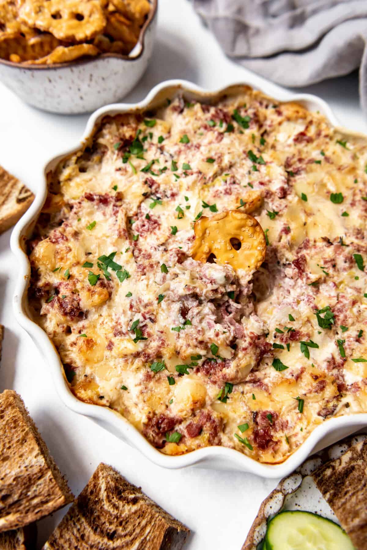 Close up of hot reuben dip in a fluted dish, with a flat pretzel wedged in the center. 