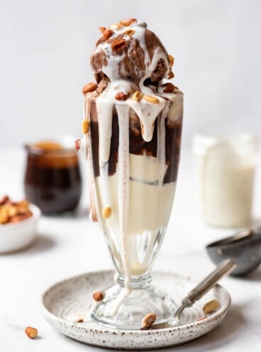 A tin roof sundae with marshmallow sauce, chocolate sauce, and peanuts in a tall glass on a plate.