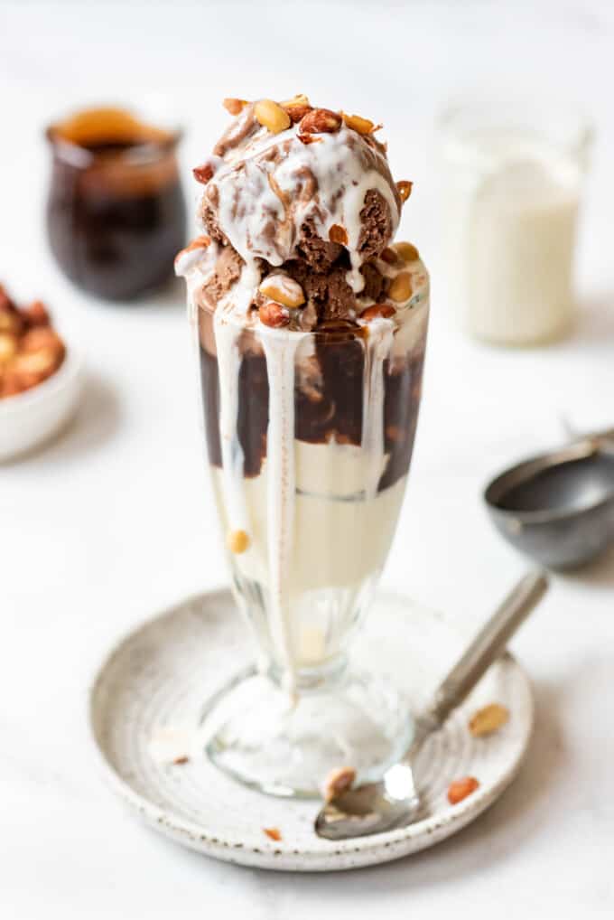 Tin roof sundaes in tall glass dessert cups with chocolate sauce and marshmallow topping behind it.
