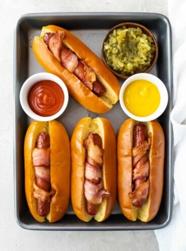 Air fryer bacon wrapped hot dogs on a small baking sheet with small bowls of ketchup, mustard, and relish.