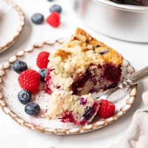 A slice of mixed berry breakfast cake on a plate with blueberries and raspberries and a bite taken out of it.