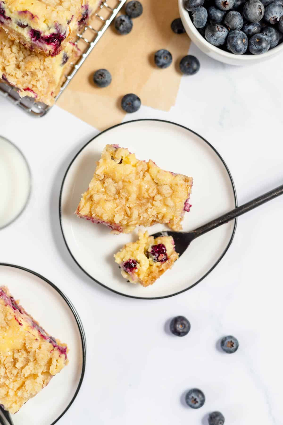 A blueberry cheesecake bar on a white plate with a fork holding a bite of the dessert.