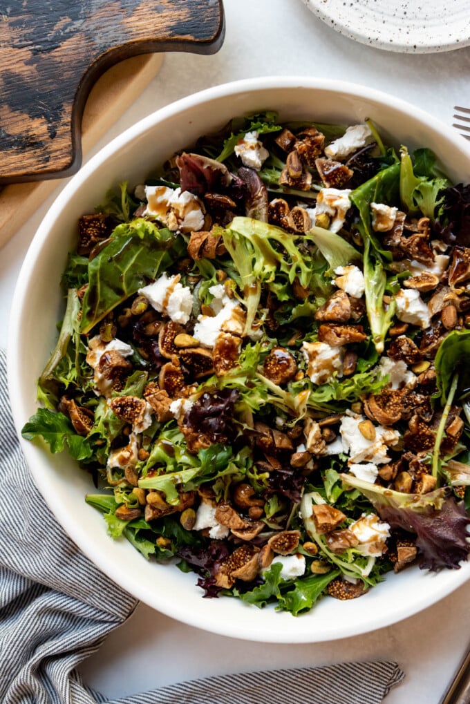An overhead image of a mixed greens salad in a large white bowl with goat cheese, pistachios, and figs.