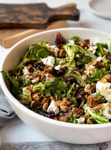 A side view of a large white bowl filled with salad with goat cheese, pistachios, and figs.