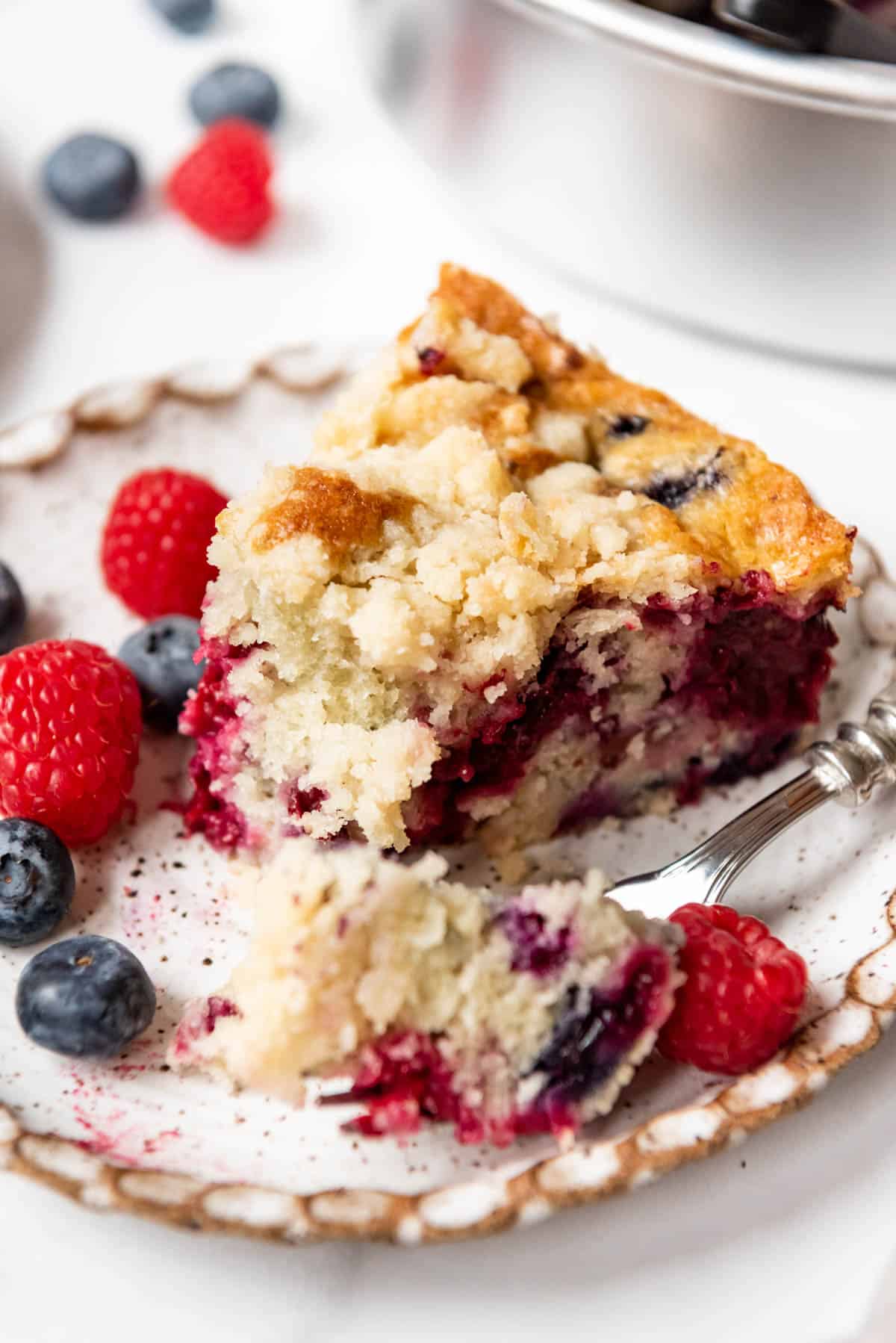 A slice of mixed berry breakfast cake on a plate with blueberries and raspberries and a bite taken out of it.