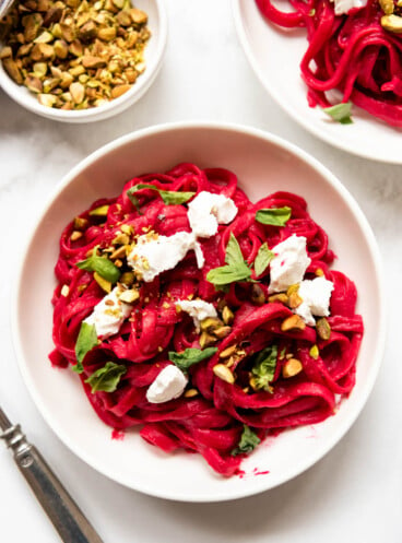 An overhead image of goat cheese, pistachios, and torn fresh basil sprinkled on top of beet pasta in a white pasta bowl.