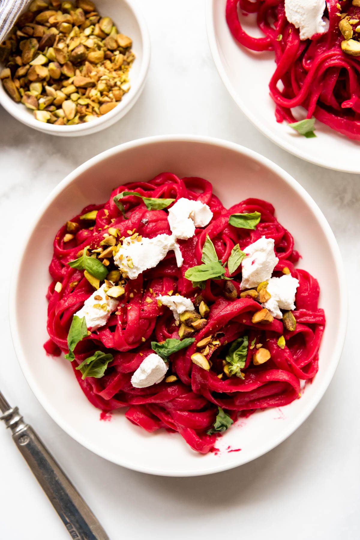 An overhead image of goat cheese, pistachios, and torn fresh basil sprinkled on top of beet pasta in a white pasta bowl.