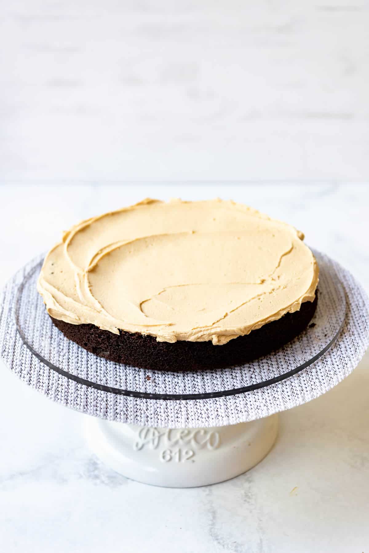 A white cake stand with a wingle tier of chocolate cake on it, with peanut butter frosting on top.