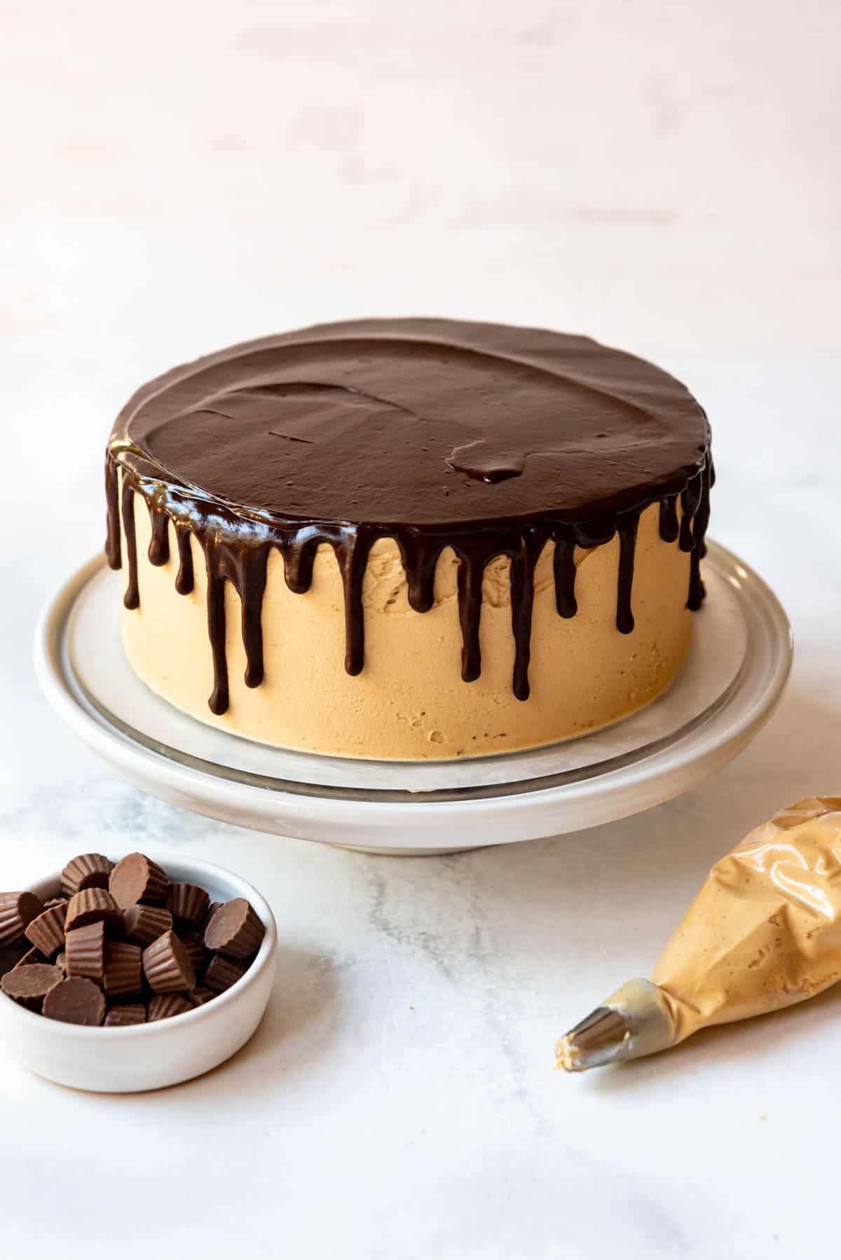 A chocolate peanut butter cake with peanut butter frosting on it, and chocolate ganache on top, dripping down the sides, on a white cake stand.