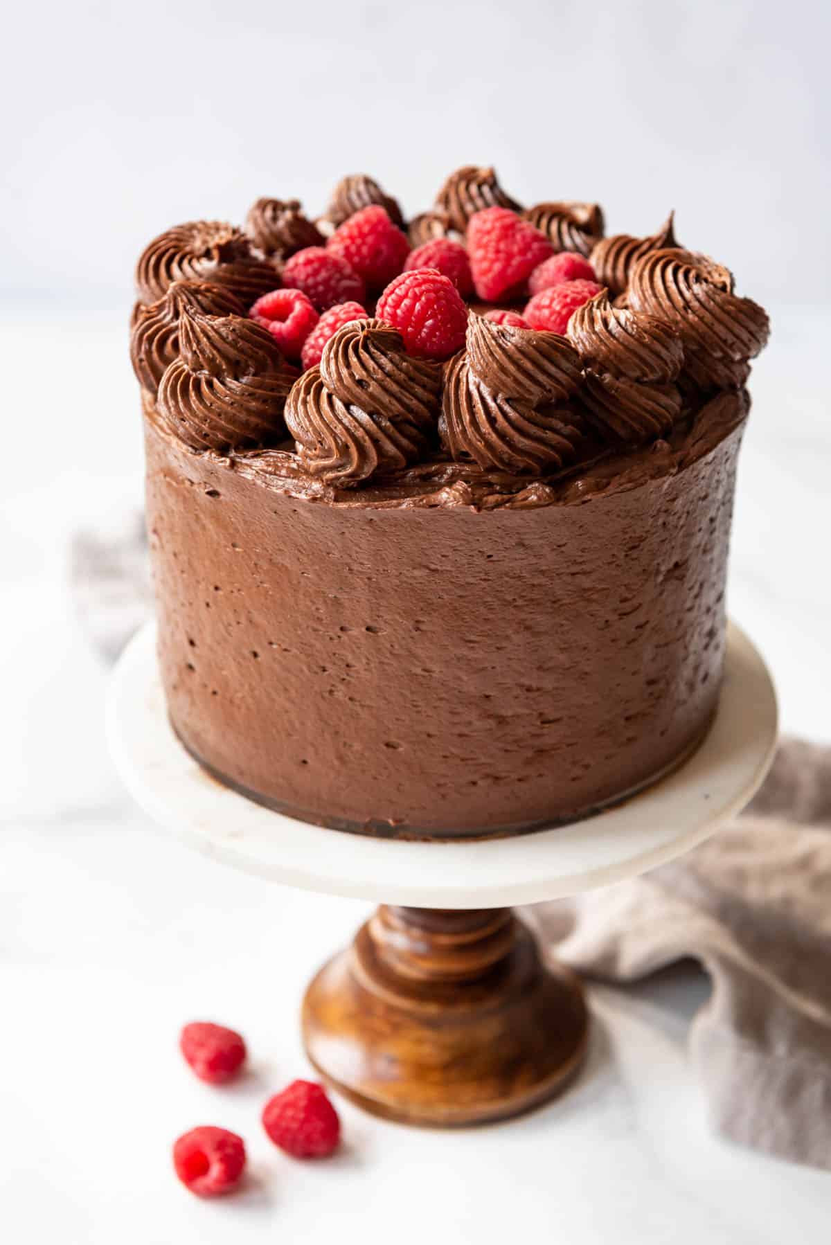 A raspberry chocolate cake decorated with fresh raspberries and swirls of homemade chocolate buttercream on top on a cake stand with three fresh raspberries below it.