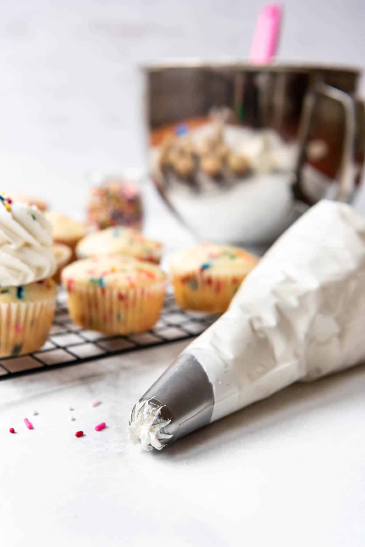 A piping bag filled with white Swiss meringue buttercream and fitted with a closed star piping tip in front of cupcakes ready for decorating.