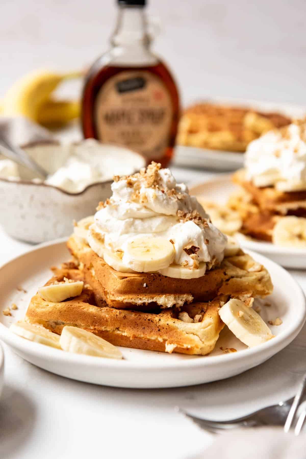 An image of banana pecan waffles on a plate with whipped cream on top and a syrup jar behind it.