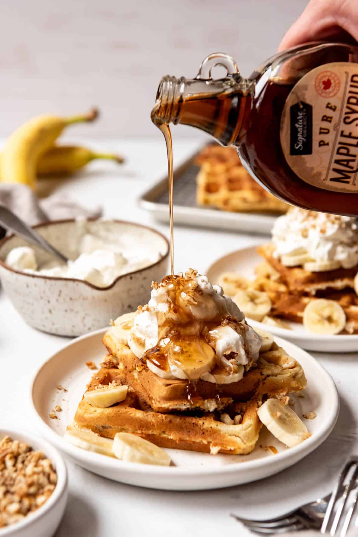 An image of maple syrup being poured over banana pecan waffles on a plate.