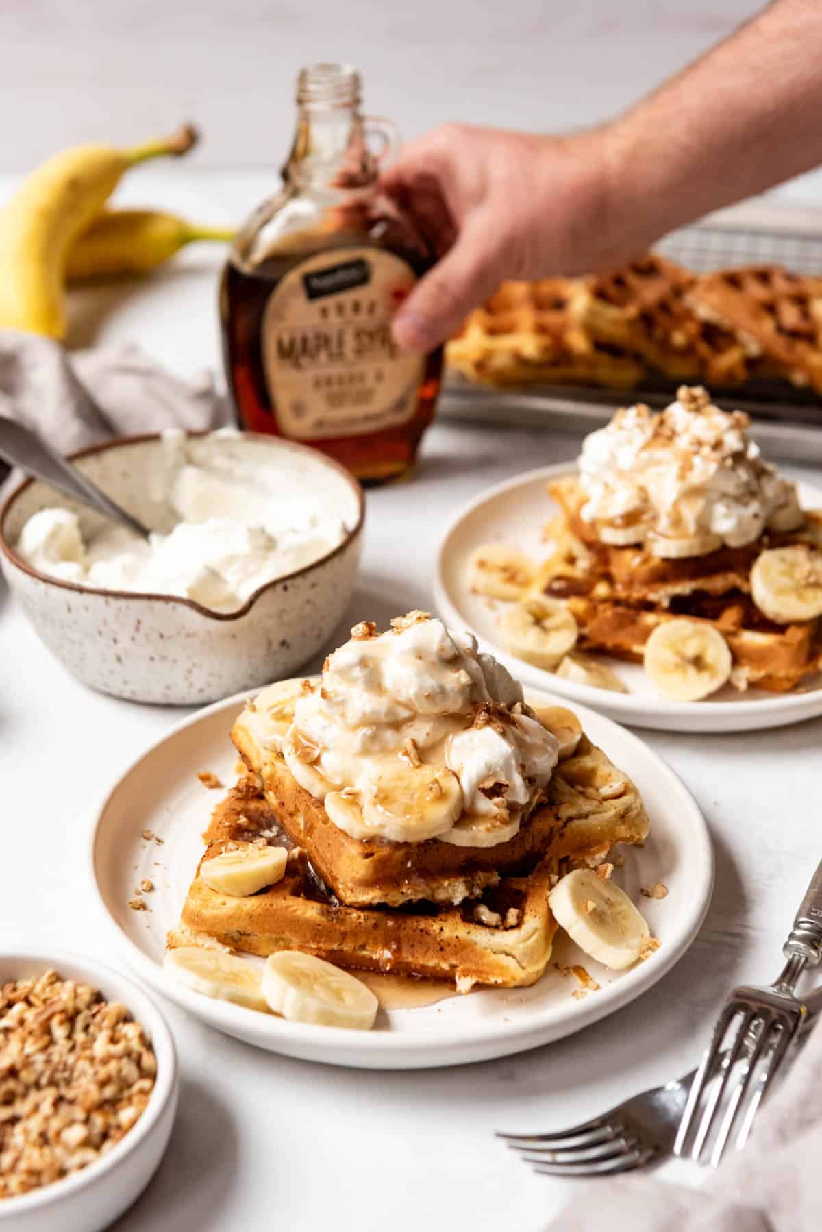 A hand reaching for maple syrup to pour over plates of banana pecan waffles.
