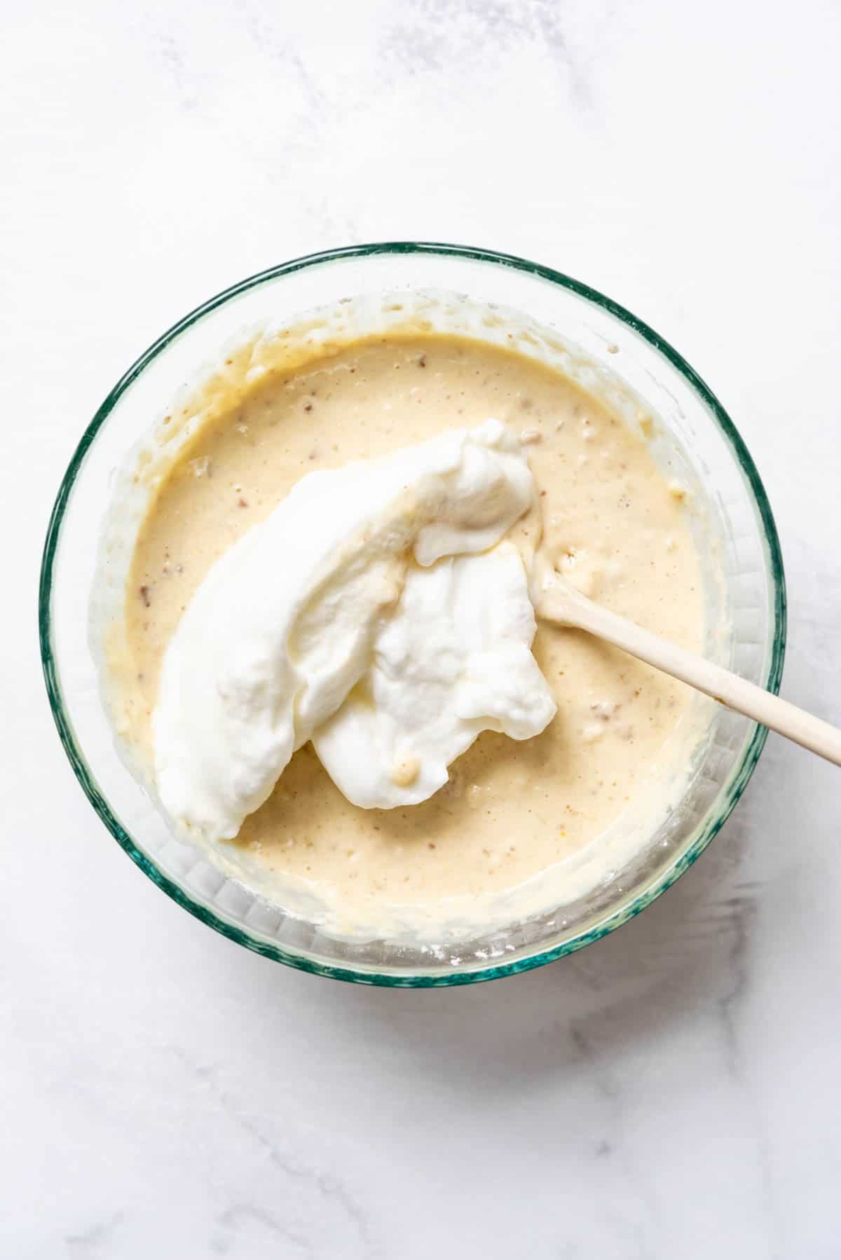 Adding egg whites that have been beaten to stiff peaks to banana pecan waffle batter in a glass mixing bowl.