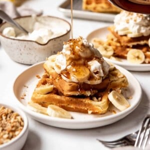An image of maple syrup being poured over banana pecan waffles on a plate.