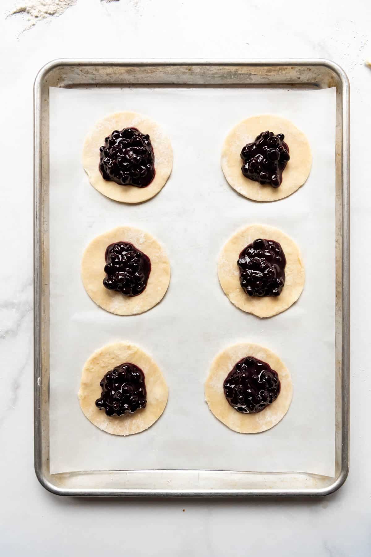 Adding blueberry pie filling to circles of pie crust on a baking sheet to make hand pies.