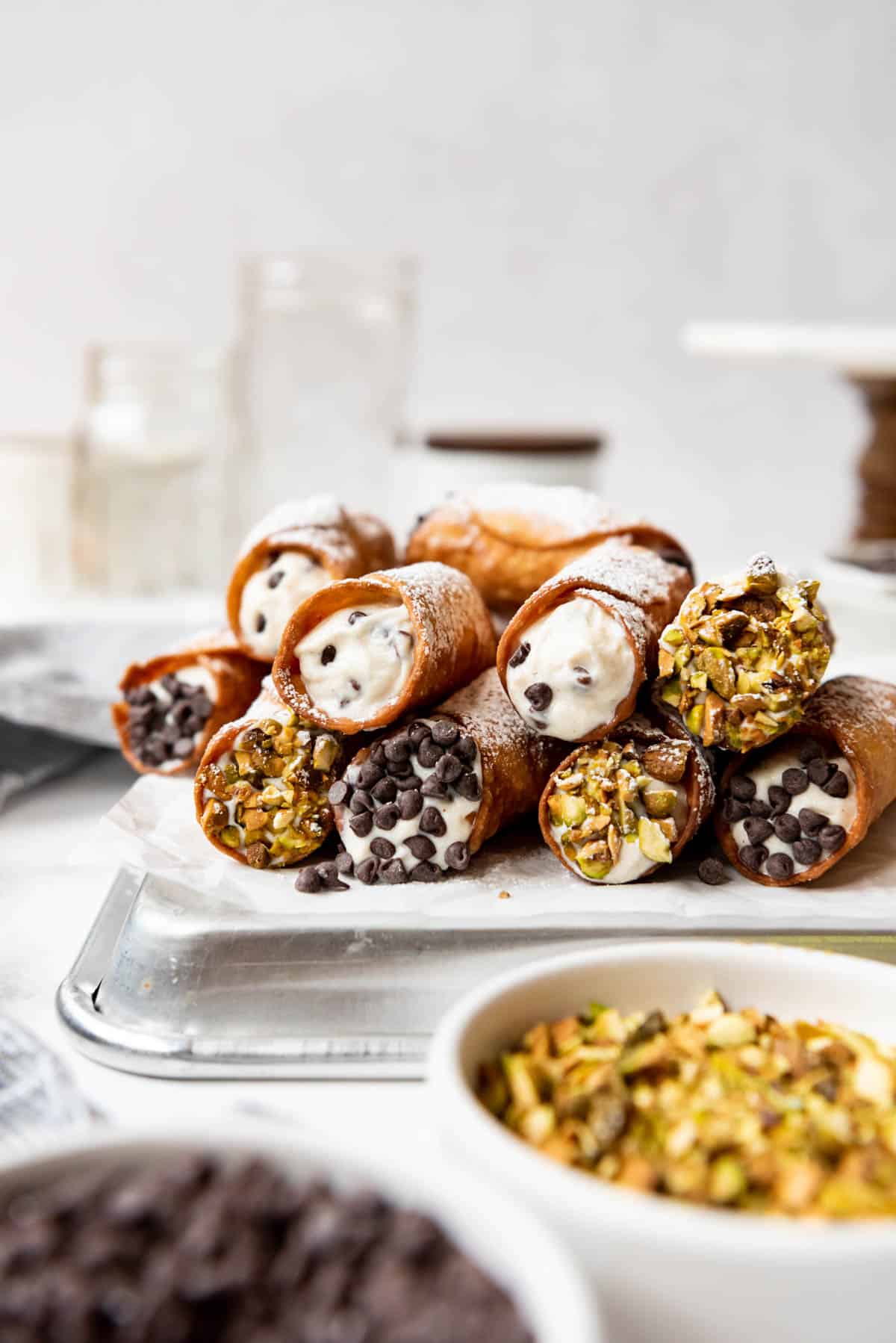 Stacks of cannoli with bowls of chopped pistachios and mini chocolate chips.