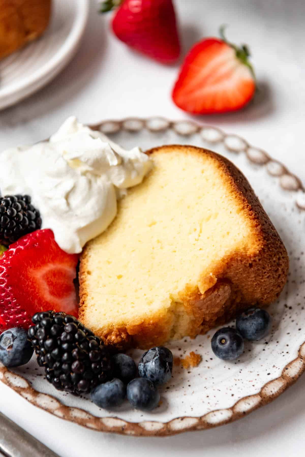 A slice of moist pound cake on a plate with fresh berries and cream.