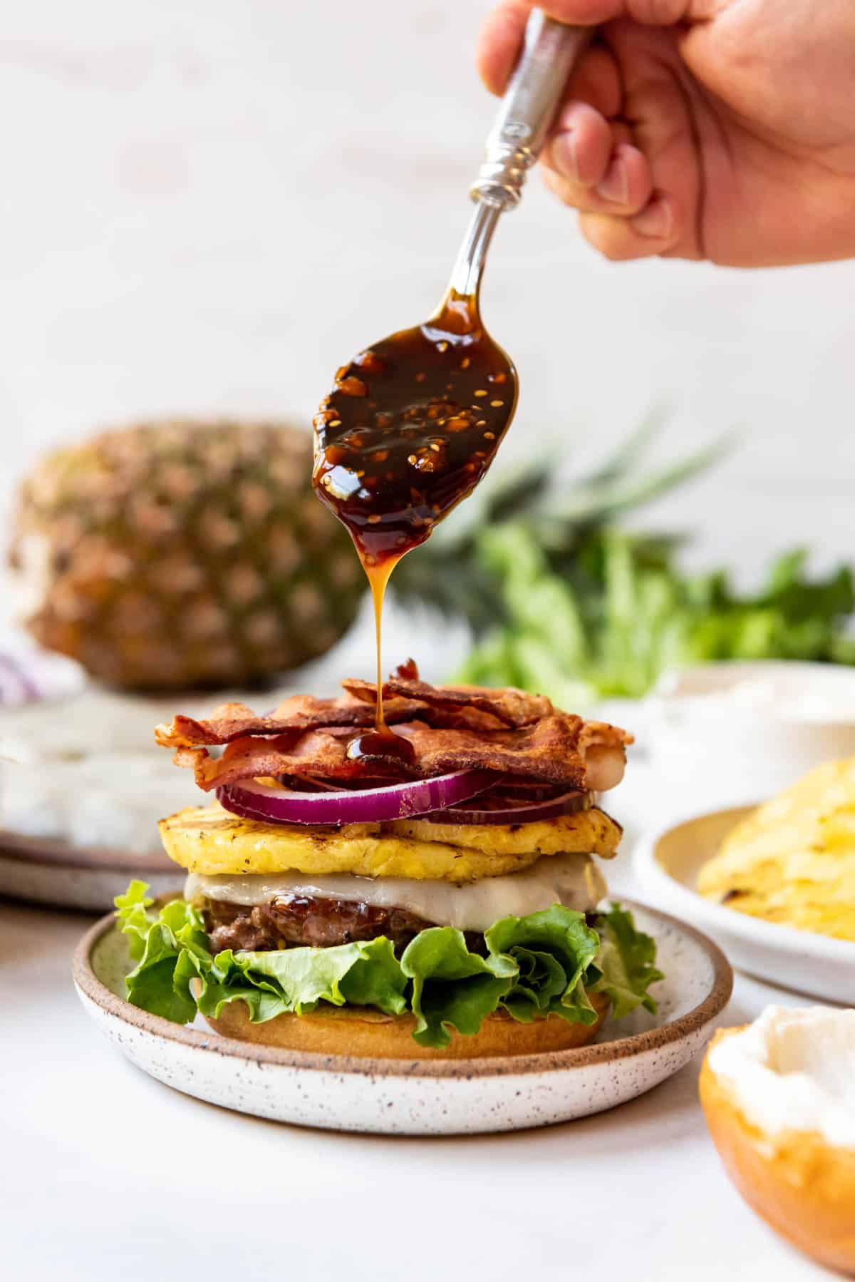 Drizzling teriyaki sauce over burgers topped with grilled pineapple, lettuce, red onions, bacon, and cheese.