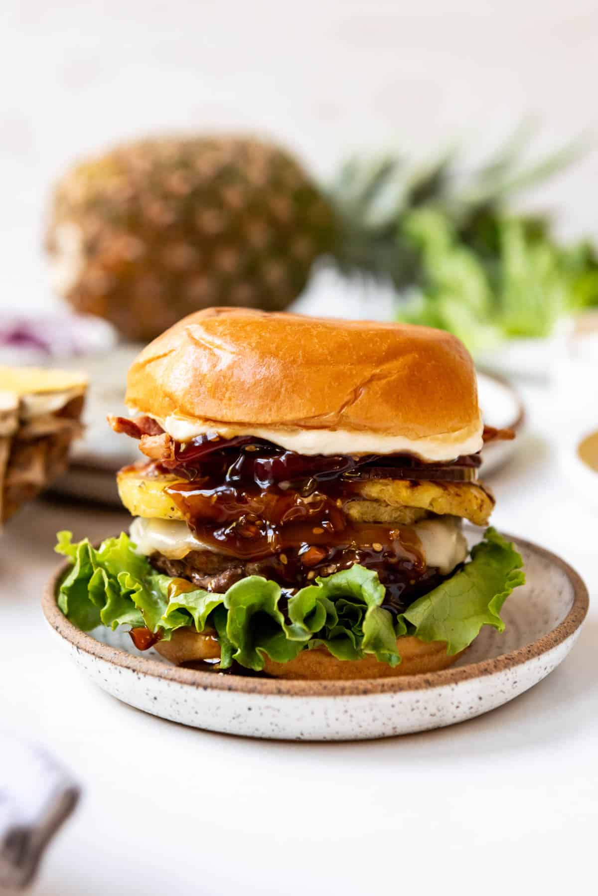 An image of a Hawaiian burger with grilled pineapple and teriyaki sauce in front of a whole pineapple.