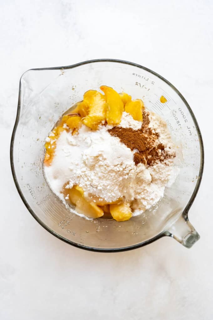 Adding sugar, flour, and spices to sliced peaches in a glass mixing bowl.
