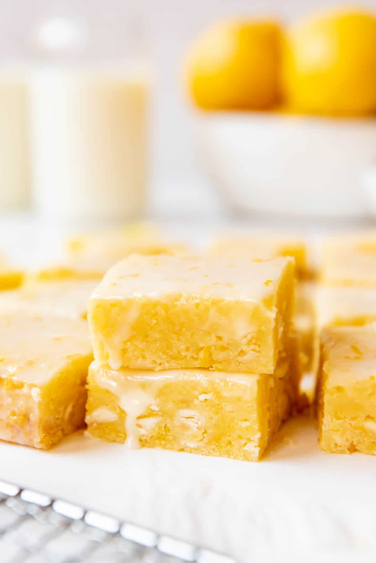 Stacked lemon brownies in front of a bowl of lemons and glasses of milk.