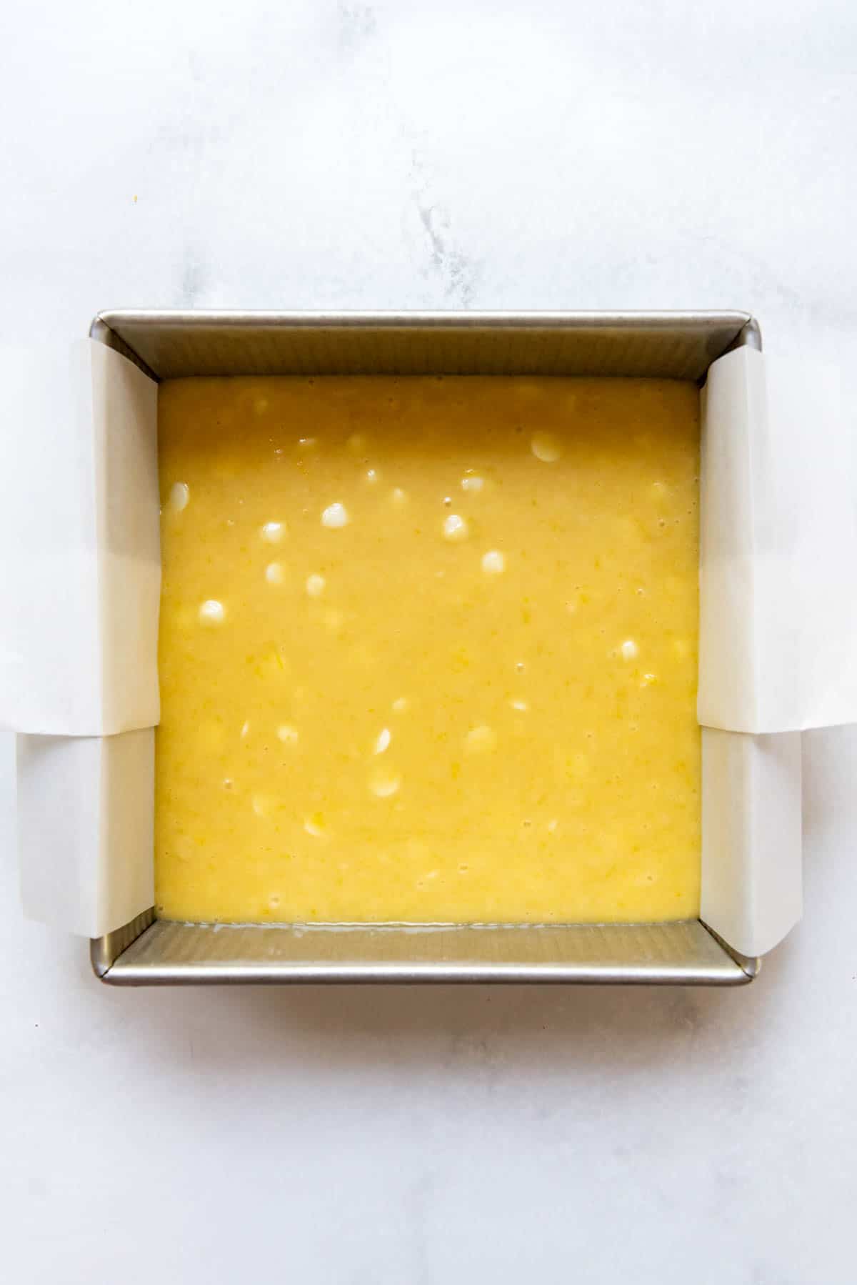 Lemon brownie batter with white chocolate chips in an 8-inch square baking pan lined with a parchment paper sling.